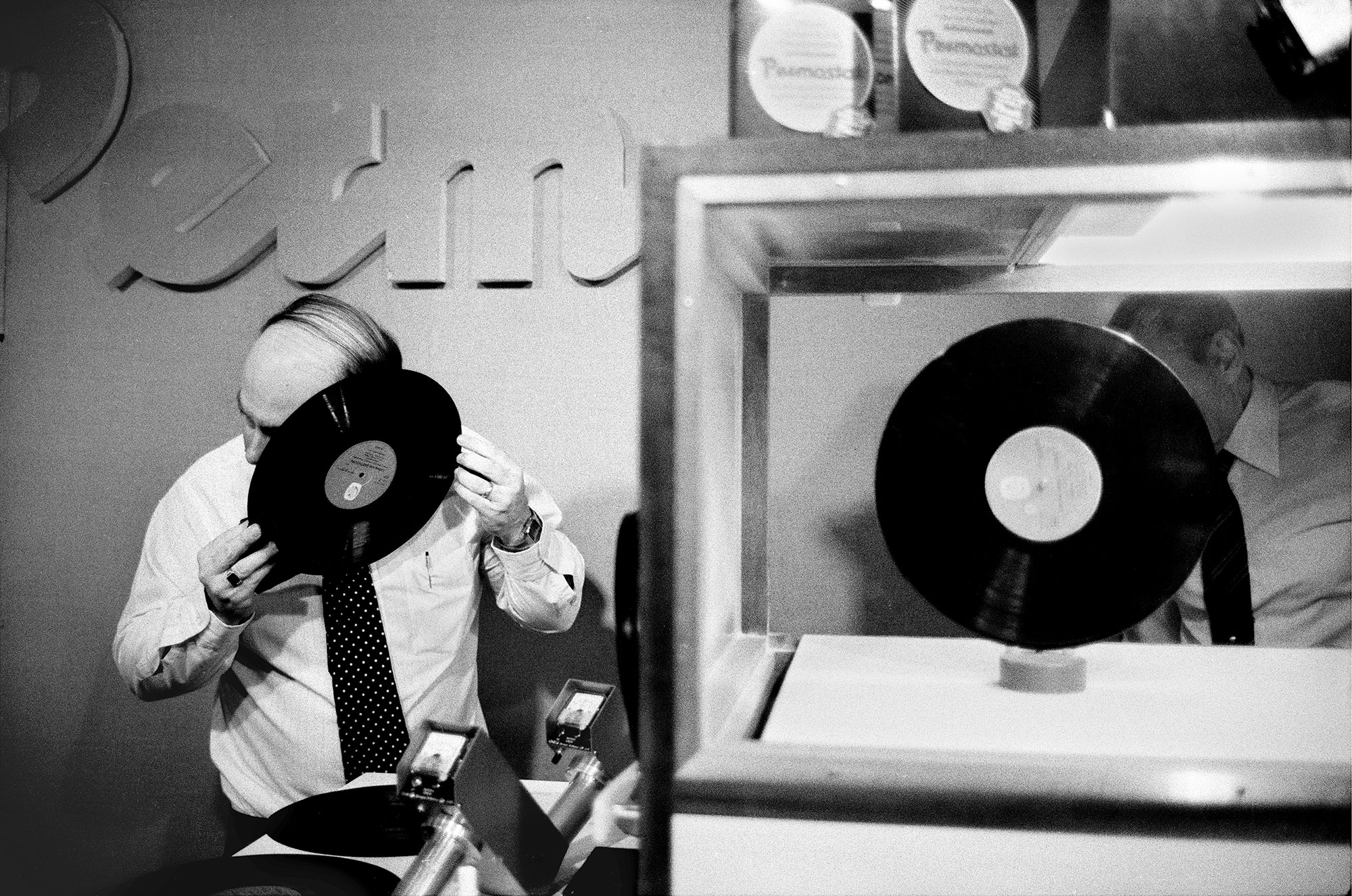 Photo horizontale en noir et blanc de Guy Le Querrec – Magnum Photos Un disque vinyl trône dans une vitrine d’exposition. À côté, un homme dégarni en chemise et cravate colle son oreille contre la galette d’un autre vinyl.