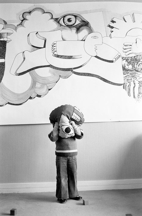 Photo verticale en noir et blanc de Jean Gaumy – Magnum Photos Un enfant en pantalon pattes d’eph’ est debout devant un mur blanc. Il se cache le visage dans un coussin décoré d’une bouche et d’un œil. En haut du mur, une grande fresque peinte représente naïvement un homme qui vole dans les nuages, surplombé par un gros œil.