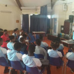 Dans une grande salle de classe aux rideaux tirés, Marie Gaumy est assise à côté d’un petit écran allumé et parle au micro devant une vingtaine de collégiens en uniforme blanc et bleu ciel.