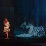 Photo horizontale en couleur de Pierre Grosbois pour le spectacle : « Dormir 100 ans » de Pauline Bureau. Dans une forêt, la nuit, une jeune fille en jupe, sweat et basket jette un œil par-dessus son épaule vers un immense tigre blanc assis comme un sphynx.