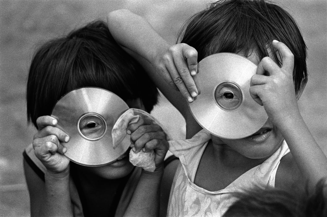 Photo horizontale en noir et blanc de Philip Jones Griffiths. Magnum Photos Deux petites filles asiatiques aux cheveux courts dissimulent chacune leur visage derrière la galette argentée d’un CD. Elles nous observent, l’œil collé contre le trou central de leur disque.