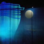Photo horizontale en couleur de Gautier Boxebeld Colonna pour le spectacle : « La révérence » de Hala Goshn Sous un ciel de pleine lune, la forme fantômatique d’un bâteau bleu et turquoise ondule sur un rideau transparent.