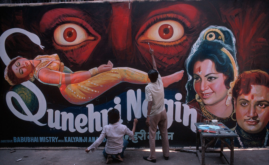 Photo horizontale en couleur de Bruno Barbey – Magnum Photos Deux hommes sont de dos, côte à côte. L’un est debout, l’autre accroupi. Ils peignent l’affiche d’un film Bollywood sur un mur qui occupe toute l’image. Une petite table en bois couverte de touches de couleurs leur sert de palette. Bras tendu en l’air, l’homme debout finalise deux immenses yeux rouges écarquillés d’effroi. Sous les yeux rouges, une jeune-femme en sari safran est étendue, la tête renversée en arrière, les yeux clos. Le titre du film : « Sunehni Nagin » épouse les courbes de la jeune femme et forme son lit. Le S majuscule est un serpent dressé menaçant la belle endormie. Le peintre accroupi pose la dernière touche de blanc sur le titre qui se termine sur les portraits de deux acteurs indiens : un homme et une femme, tous deux couverts de bijoux dorés, regards charbonneux et bindi sur le front.