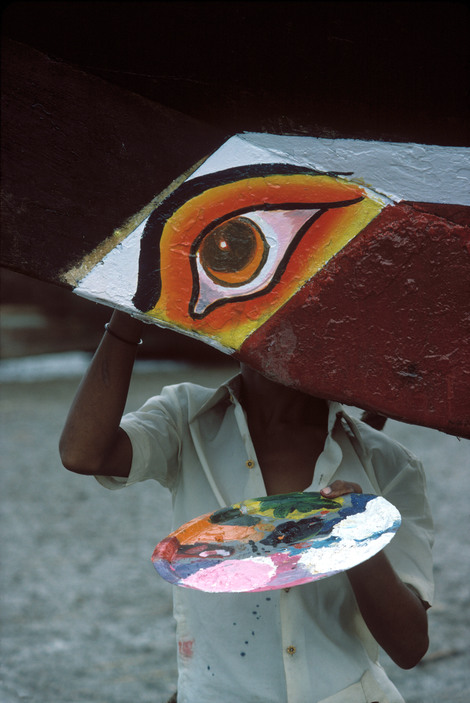 Photo verticale en couleur de Bruno Barbey – Magnum Photos Un œil au sourcil noir, à la paupière fardée de jaune et orange est peint sur une poutrelle qui barre la moitié supérieure de la photographie. La tête cachée par la poutrelle, un homme en chemise blanche se tient debout juste derrière l’œil qui lui fait office de visage. Il tient une palette pleine de couleurs dans la main gauche. Il nous fixe de son œil cyclopéen.