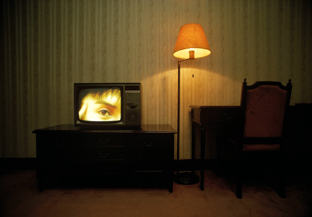 Photo horizontale en couleur de René Burri – Magnum Photos. Un œil nous fixe sur l’écran d’un gros poste de télévision allumé. Le poste repose sur un meuble bas et sombre. À côté, un bureau en bois laqués tout aussi sombre. Entre deux, une lampe à pied et son abat-jour ocre éclaire faiblement le papier peint vieillot d’une chambre moquettée de marron.